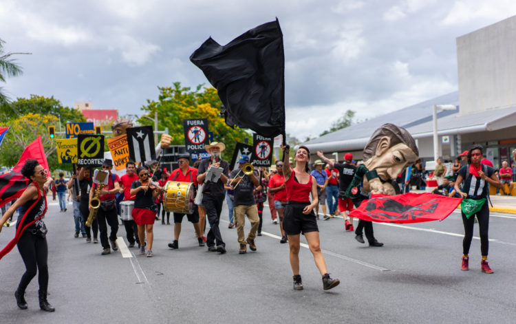 Papel Machete in the streets for the May 1st march in San Juan, Puerto Rico. At the center-front of the image, a woman in a red tank top and black shorts waves a large black flag. She is flanked by two other woman in black, waving large red flags with the word 
