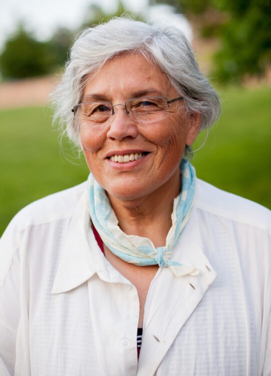 Filmmaker Lourdes Portillo, a woman with short gray hair, glasses, a white shirt and a blue and white scarf around her neck, smiles at the camera outdoors with greenery in the background.