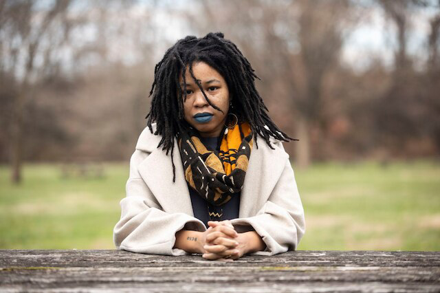 A brown-skinned Black person with shoulder length locs and bright blue lipstick is sitting at a wooden picnic table wearing a tan-colored coat with a mudcloth patterned scarf around their neck. Behind them is a park landscape with grass, overcast sky, and bare trees in late winter.