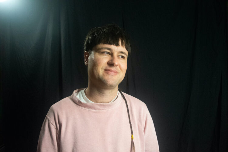 A Pākehā (white person from Aotearoa New Zealand) with a brown-haired bowl cut and a braided rats tale wearing a soft pink sweatshirt with a baggy collar standing in from of a black curtain, mildly smiling into space just off-camera.