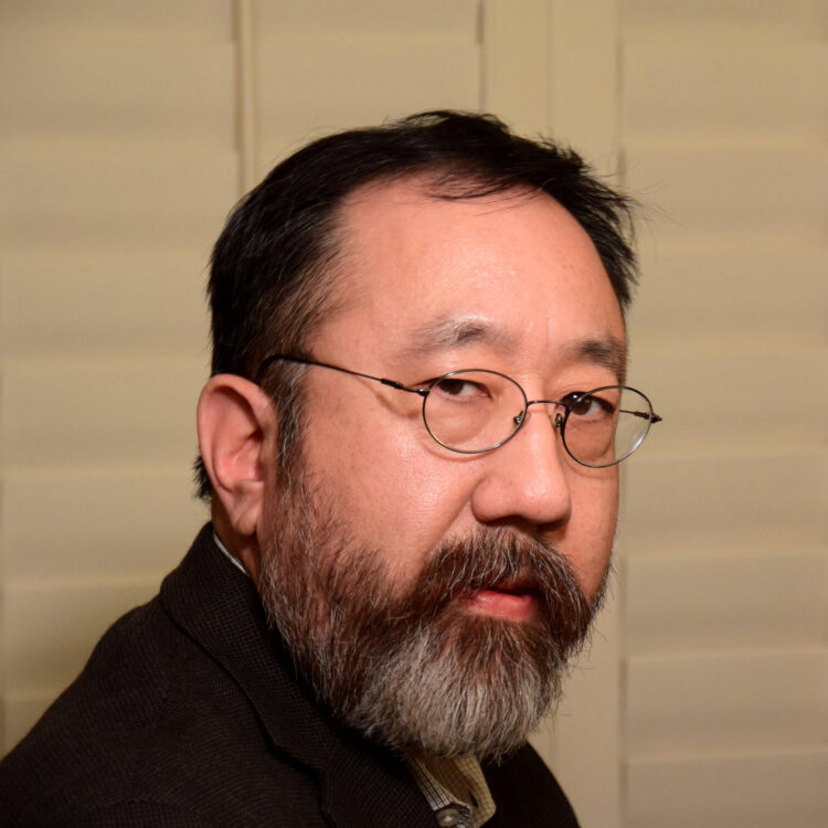 An Asian man with short black hair, beard, and moustache, wearing glasses, a dark jacket, and a finely checkered shirt. Behind him are large white shutter panels.