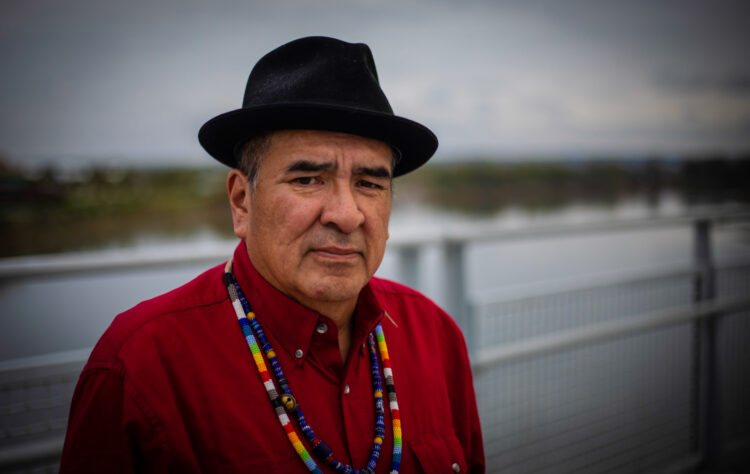 An indigenous man with short black hair, wearhing a black fedora, red button down shirt, and two multi-color beaded necklaces stands in front of a railing above the Missouri River.