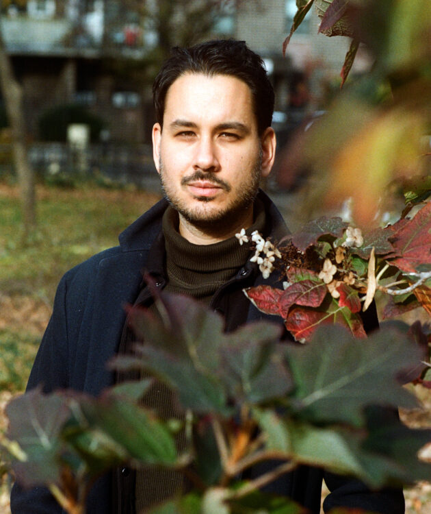 A light-skinned man of South Asian descent with dark black hair, a short scruffy beard, dressed in a green turtleneck with black shirt and blue dress coat surrounded by flowers.