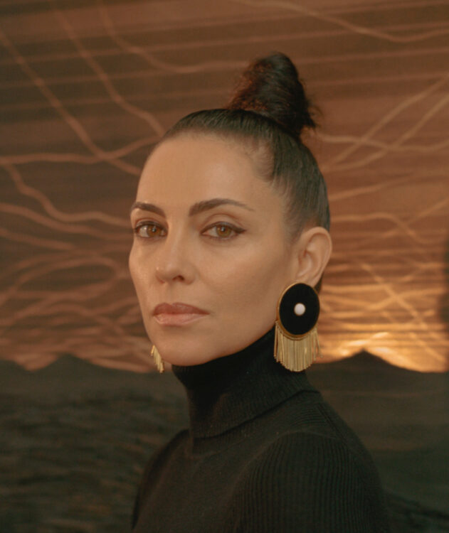 A Latina woman with dark hair in a high bun, wearing a black turtleneck and large black and gold earrings. She is in front of an artwork panel that is golden, metallic, and black.
