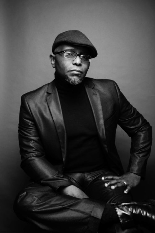 Tyehimba Jess in Black and White photo with cap on wearing suit. No smile.