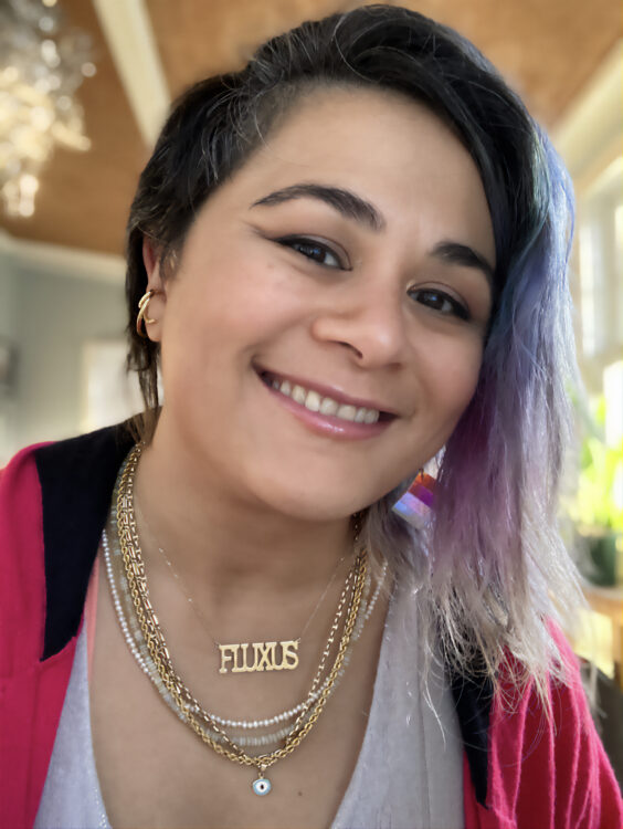 Photo of artist with asymetrical hair, short black on left side and longer faded purple on right. She has tan skin and is smiling. She is wearing a pink hooded sweater with gold jewelery. Her necklace nameplate reads Fluxus.
