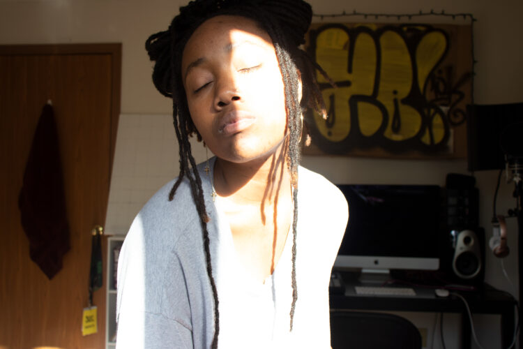 A Black Woman with locs in a bun, wearing a light grey shirt, sits in a room with her eyes closed and head tilted to the right. Behind her is a bedroom with a studio setup and yellow graffiti wall art.