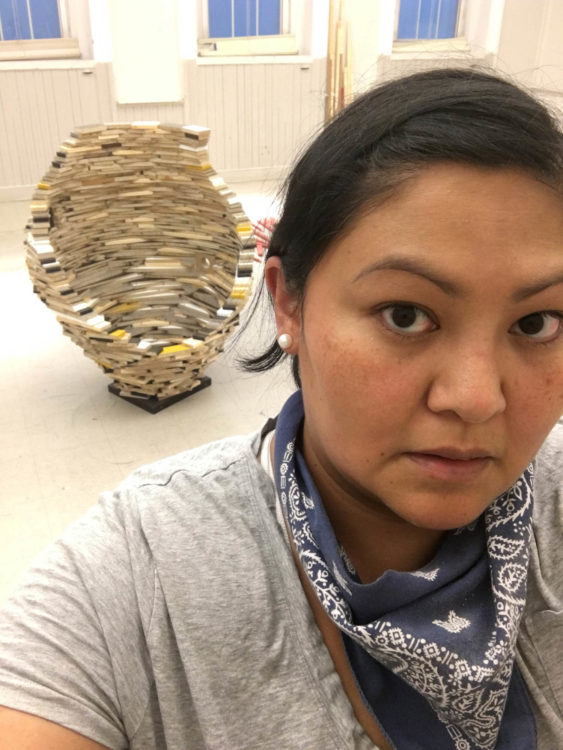 A Native American woman is standing in an art studio wearing a navy blue bandana, a grey shirt, and pearl earrings. Behind her is an abstract sculpture made out of IKEA remnants. 