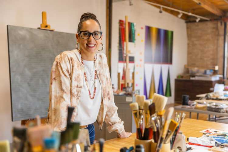 Headshot photo of an artist standing in her art studio, smiling with both hands wresting on a work table in front of her.