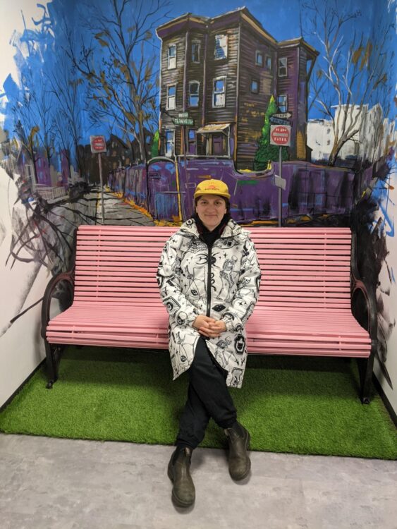 A white person in a white jacket and yellow hat sits on a pink bench with her hands folded in her lap and her legs crossed. Behind her is a mural of a house painted on the wall.