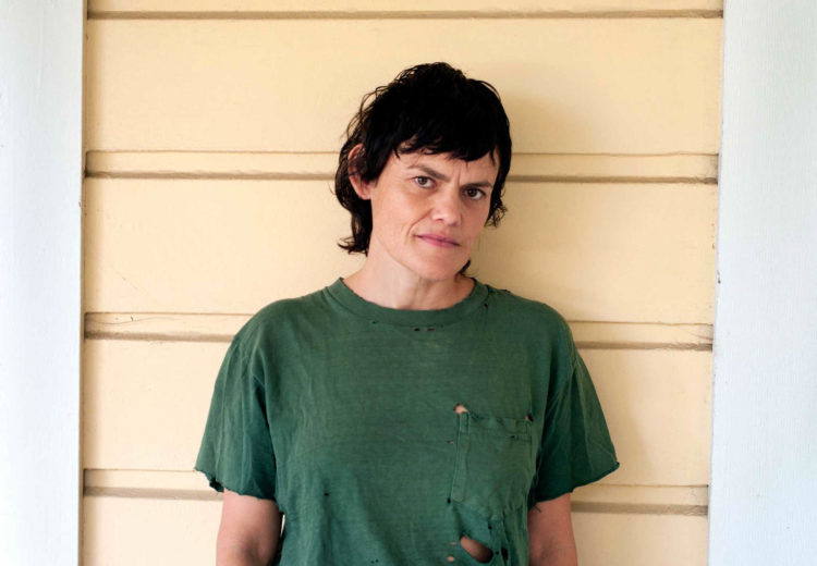 Lesbian with brown hair and old green shirt leans against yellow house.