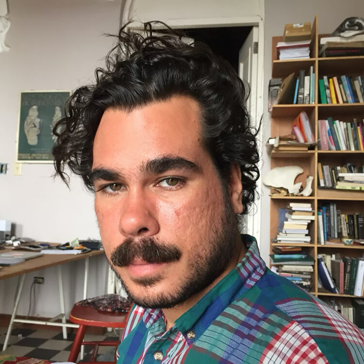 Portrait of Ramón Miranda Beltrán with disheveled black curly hair, a mustache and short facial hair, green eyes wearing a green, blue, red and white plaid button down shirt. Behind him a bookshelf filled with books, folders and various bones on his right side. On his left side a disorganized desk with various computer hardware. On the wall a three color screen print poster of an archaeological exhibition depicting a front and side view of a Puerto Rican indigenous carve stone.