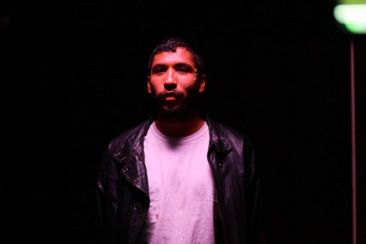 A brown-skinned person standing wearing a black leather jacket and white t-shirt underneath, smiles slightly. They stand under a light purple red hued light and look forward past the camera prepared, confident, and ready to take their next step. In the upper right hand corner, there is a greenish white light that is part of the video shoot set.