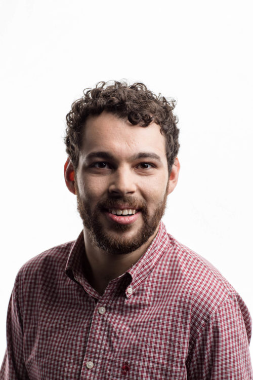 A white man with a brown beard and short curly hair subtly smiles into the camera. He's wearing a red-checked button-down and is in front of a background of white.