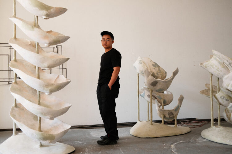 An East Asian person standing in the center of a studio, surrounded by his biomorphic sculptures.