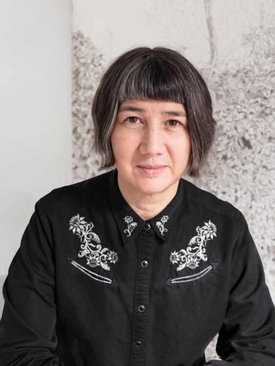 Half-Japanese, half-White woman with short dark hair wearing a black button-up shirt with silver designs, in front of a blurred gray and white background.