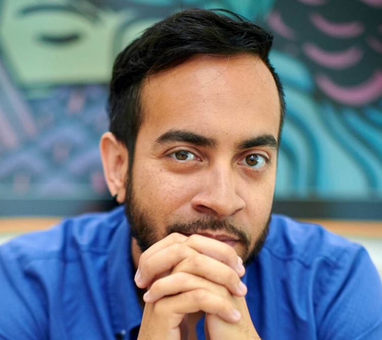 A Puerto Rican-American man wearing a blue collared shirt, leans forward on a couch with clasped hands at his chin.