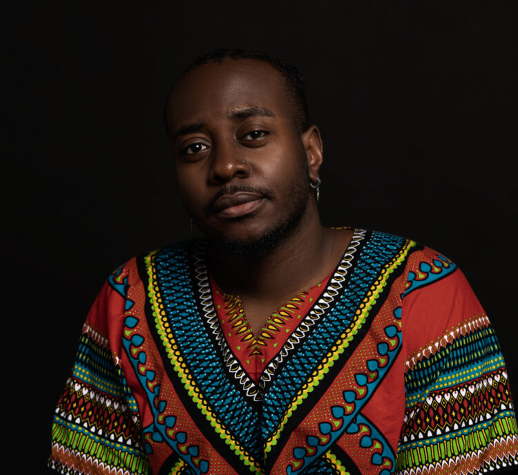 A Black man, seated on a brown stool, with short black hair braided to the back of his head. He is wearing a dashiki, multi-colored with red, blue, green, yellow, and white. On his left wrist is a brown fur necklace. On his right wrist is a silver Casio watch. He has dark blue pants on. Behind him is blackness.