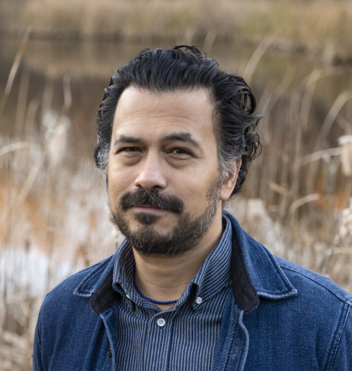 Man with black slicked back hair with a thick mustache and close cropped beard. He is wearing a striped indigo shirt, lapis lazuli beaded necklace, and denim chore coat. A pond surrounded by reeds is visible in the background.