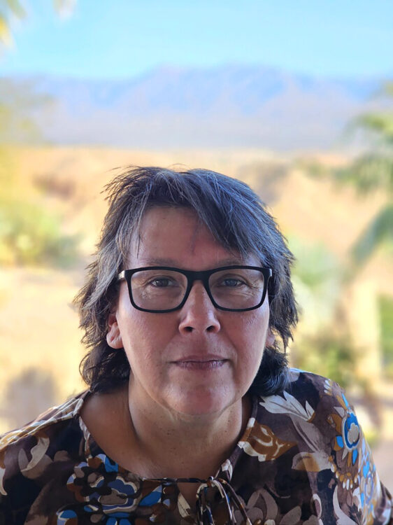 Indigenous woman with black rimmed glasses and short black and white hair takes headshot with the blurred mountains in the background