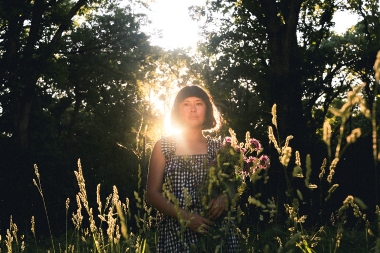 Woman with a dark chin length bob wearing a black and white checked sleeveless dress is backlit by the setting sun. She is standing in a wooded area, and in the foreground are wisps of tall grasses. She is holding wildflowers.