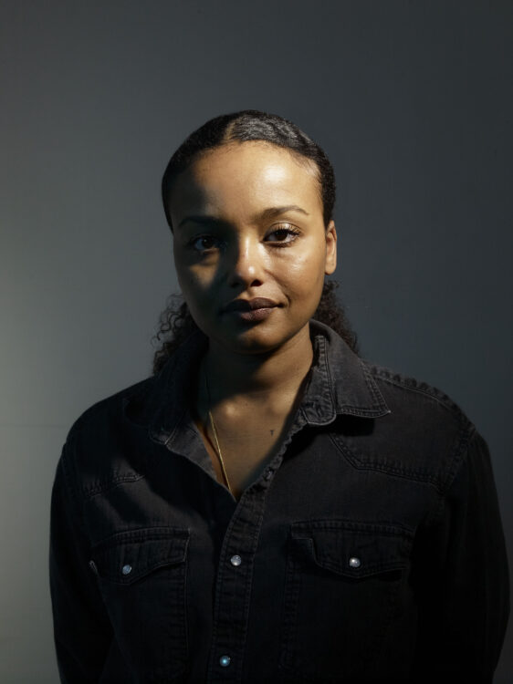 A brown skin woman wearing a black denim shirt. Her face is half cast in shadow and her dark hair curly is in a low ponytail with a middle part.
