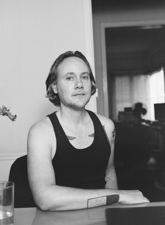 A white transmasculine person with some tattoos wearing a black tank top and sitting with his arm resting on a table.