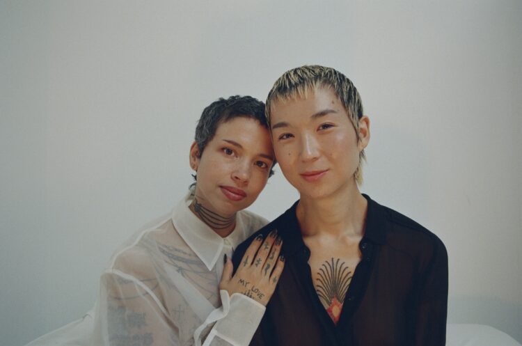 Holland Andrews and yuniya edi kwon look into the camera in front of a white-blue background. Their heads are touching and Holland's hand rests on yuniya's shoulder.