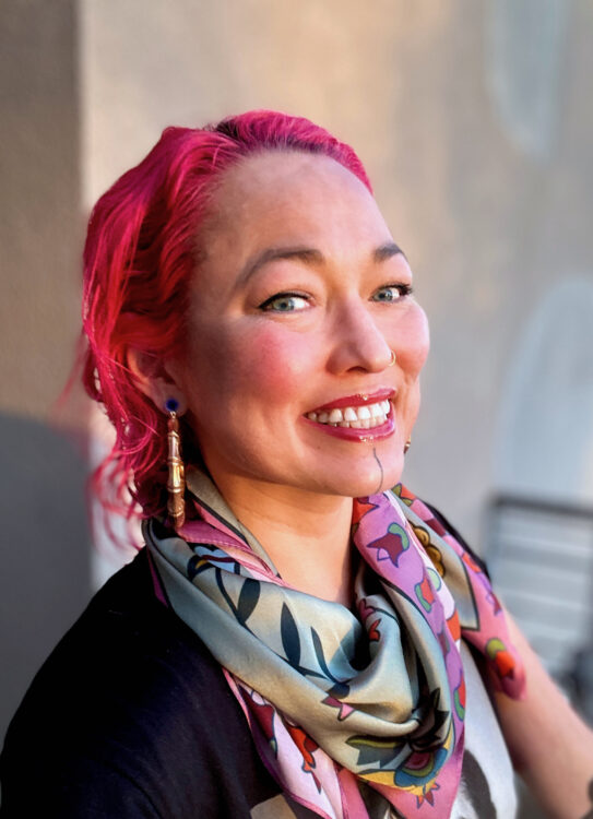 Erica Lord, a mixed-race Alaska Native woman, smiling with pink hair and gold hoop earrings