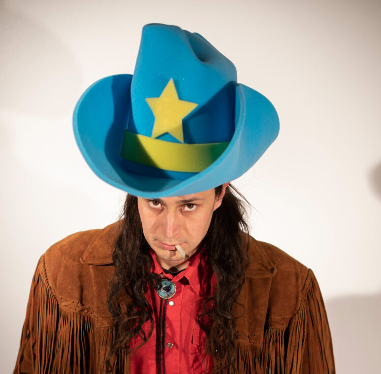 A photo of a Native American man in a large blue foam cowboy hat and red shirt and brown fringe leather jacket sitting down smoking a joint facing the camera with a gray background.