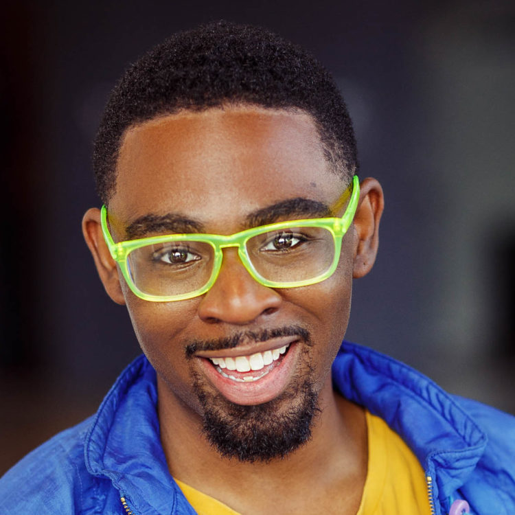 A Black man with short black hair, wearing neon green glasses a yellow t-shirt and light blue jacket. 