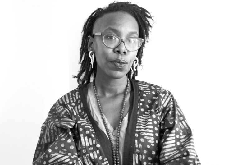 A person with deep brown skin and locs wearing glasses and a patterned loose top looks towards the camera with a soft smile.