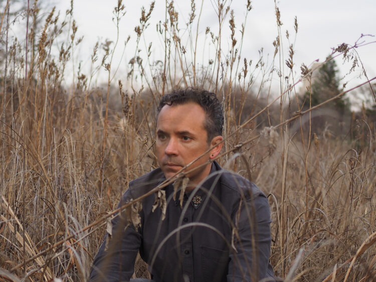 Artist Gavin Kroeber staring into the distance while sitting in a field of native prairie plants on a gray winter day.