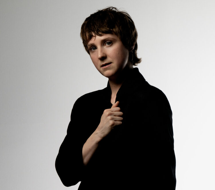 A white person with a short, reddish mullet stands before a gray-white background, looking into the camera while holding the collar of their black suit.