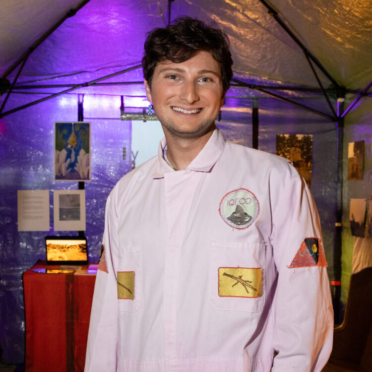 Lee stands in front of a display of past projects, wearing an orchid-colored jumpsuit for the Institute of Queer Ecology. He has fair skin, brown hair, and a big smile.