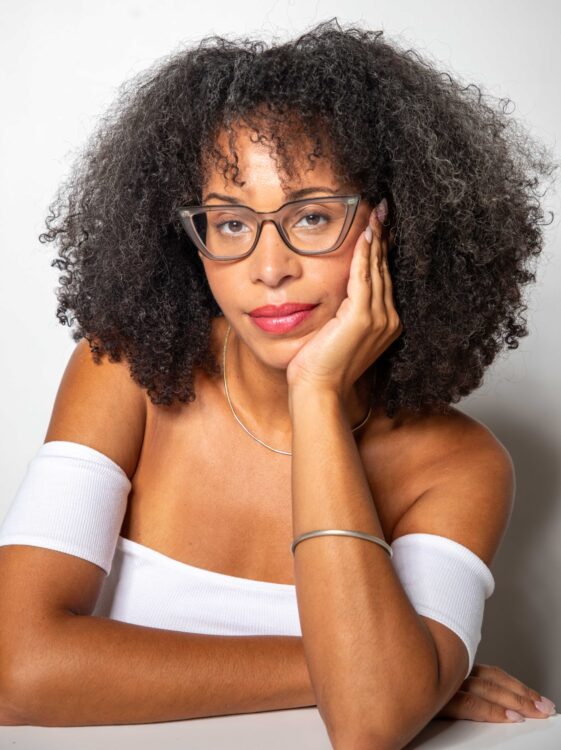 Artist Loira Limbal is a Black woman with an Afro sitting in front of a white wall. She is wearing a white blouse and gray glasses.