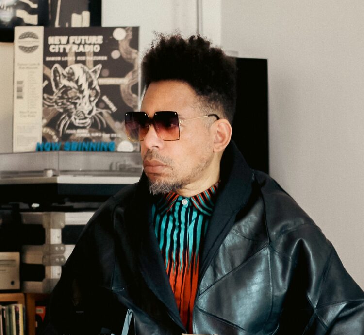 Light skinned Black man with square sunglasses and hair cropped short on the sides and back. He is wearing a brightly colored blue and red top with a black leather wrap. He is sitting amongst records. 