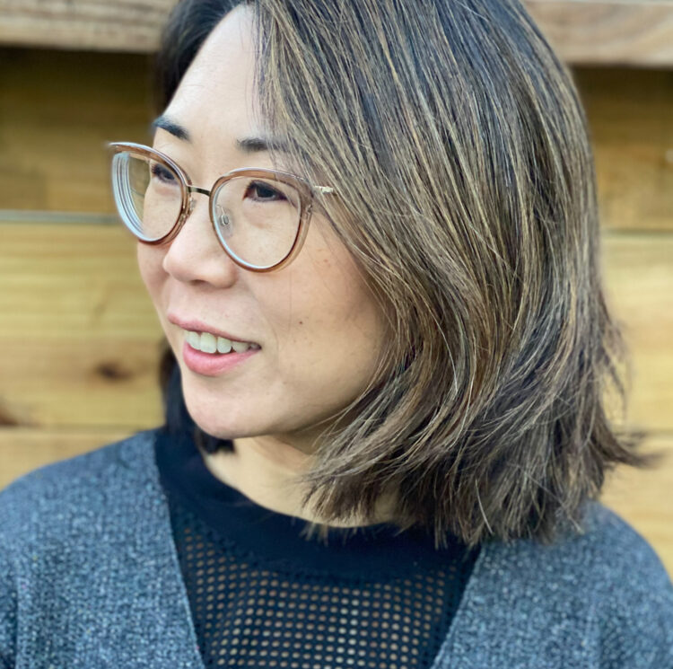A smiling asian woman with clear-rimmed glasses, shoulder-length brown hair, and wearing a black and gray sweatshirt, looks to her right in front of a wooden fence.