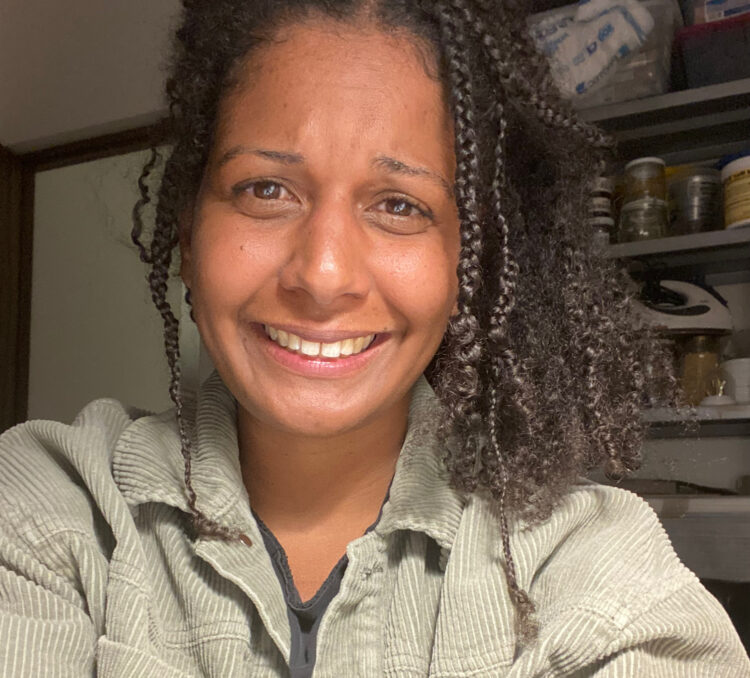 A smiling girl showing her teeth with curly hair and braids in front of her face. Reddish brown skin. Wearing a black t-shirt and olive green jacket. In the background, on the right side, there is a metal shelf with various jars and on the left side, an open door.