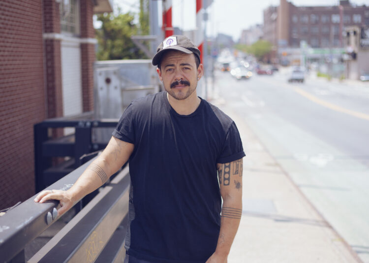 Angelo Madsen is standing on a bridge in New York. He is wearing a black t-shirt, black cap, and has light tan skin with tattoos, piercings, and a mustache.