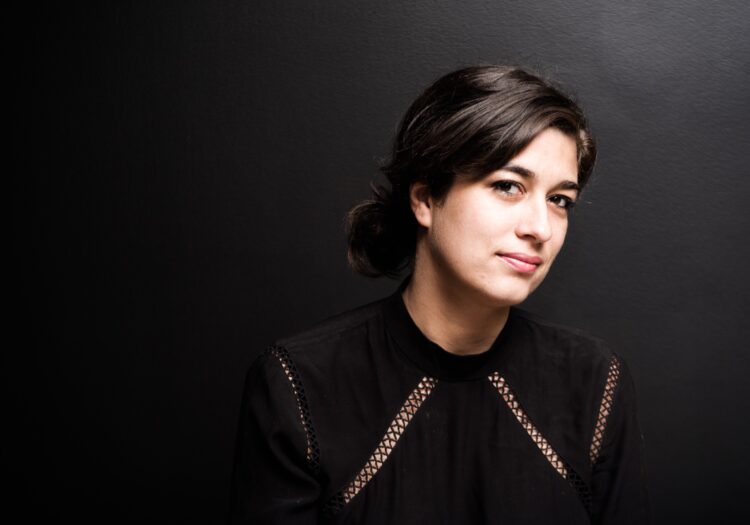 This picture is a headshot of theatre artist Marike Splint, who has dark brown hair, brown eyes eyes and olive skin. Her head is somewhat titled and she is looking directly at the camera. She's wearing a black top with mesh detailing. 