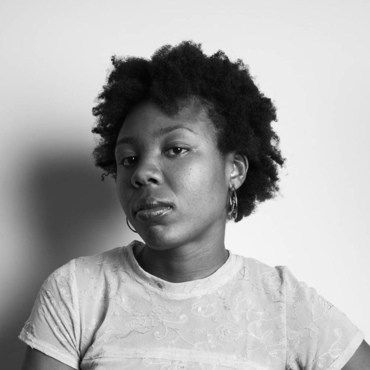 A black woman with short hair stands with her hands folded on one hip and looks at the camera.