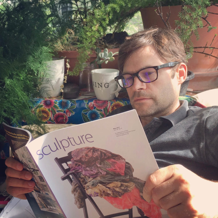 A man wearing glasses reads Sculpture magazine in a light filled sunroom surrounded by plants.