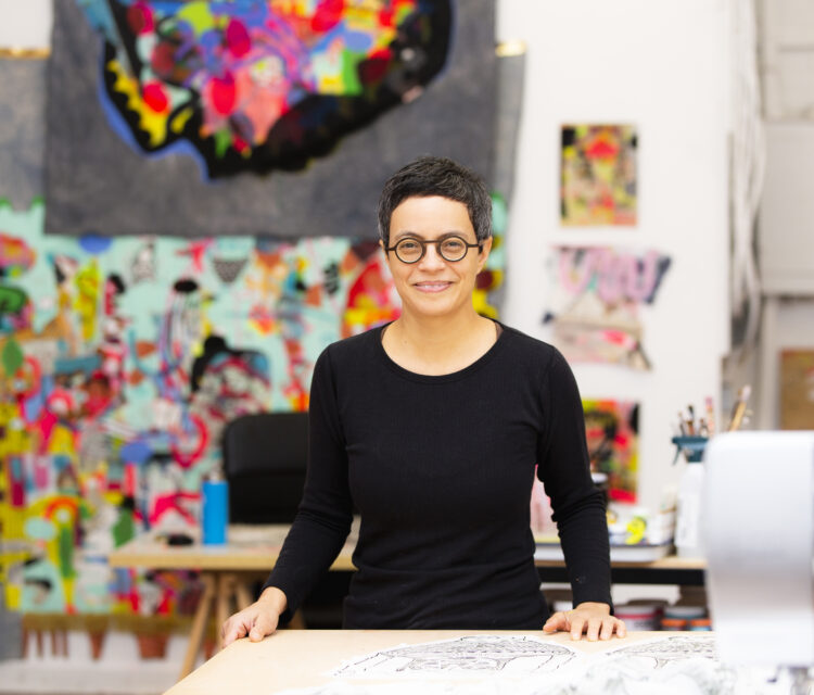 Artist with short hair and black shirt standing in the foreground with her hands on a table. The background is out of focus with colorful artworks on the wall.
