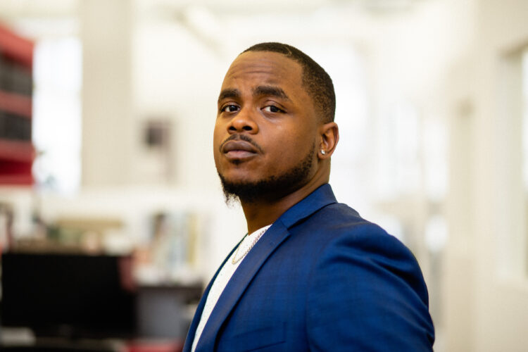 The author, an African American man, stands sideways with his head turned looking into the camera. He has short black hair, a goatee, and is wearing a blue blazer with a white crew shirt underneath and a silver Cuban link chain.