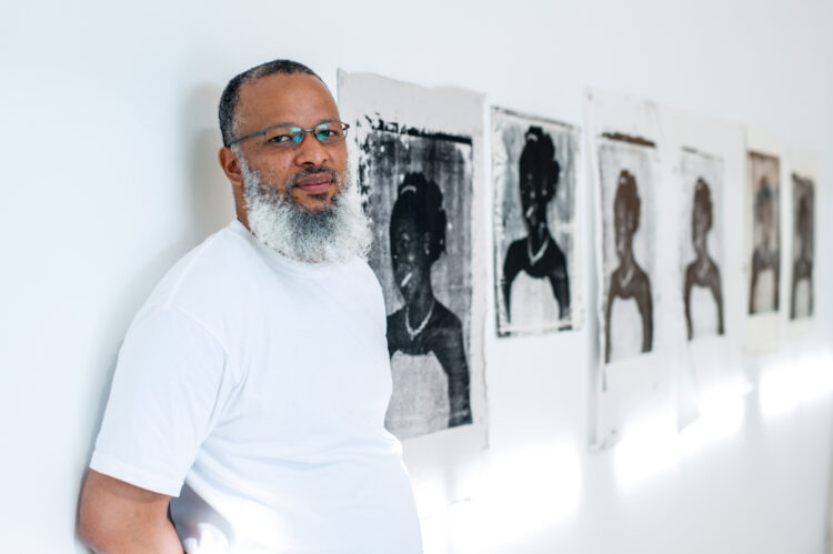 Man with white beard, wearing glasses and a white t-shirt, leaning on a wall next to 6 screen prints tests.