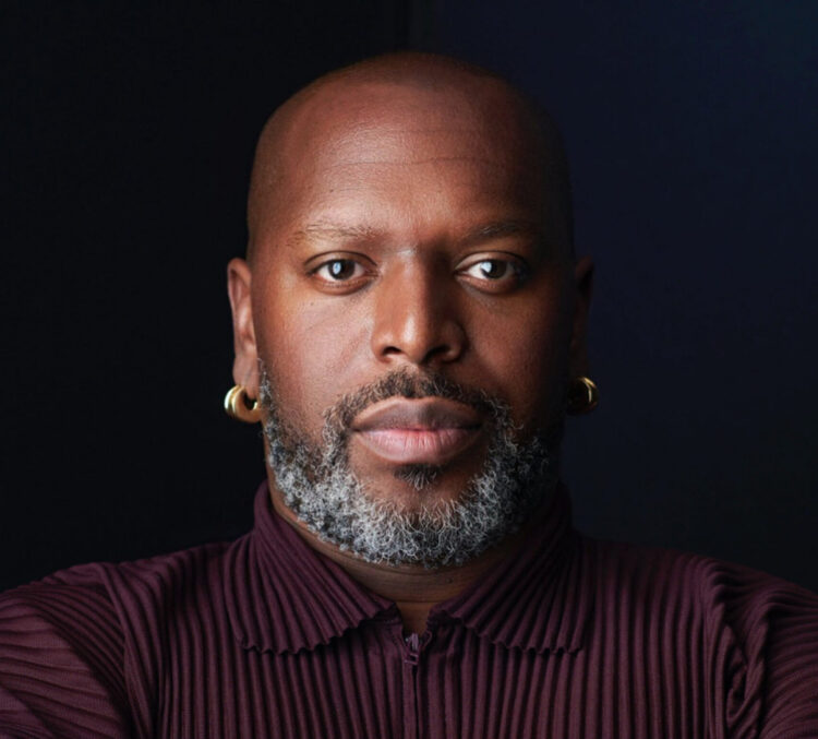 Bald African American man with a beard in a burgundy shirt and gold earrings.