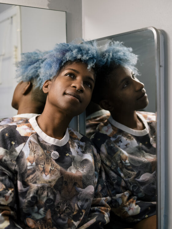 Tchaiko, blue afro with shaved sides and back is in front of a mirror that makes her appear as a three headed creature wearing a sweatshirt covered with cats