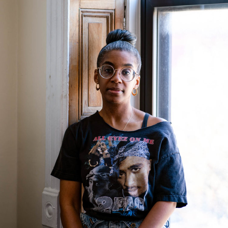A black woman with salt and pepper hair piled in a bun, wearing large, round wire-rimmed glasses and a Tupac Shakur t-shirt sits in a window framed by pocket shutters, looking directly at the camera