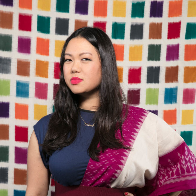 Asian American woman with long black hair wearing a navy dress with a fuschia dupatta over one shoulder. She has a hand on her waist and looks into the camera with a cloth background of handwoven multicolor rectangles. 